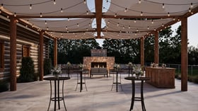 Covered outdoor patio at Casa del Rio Fresno with string lights, rustic wooden posts, stone fireplace, high-top cocktail tables with floral arrangements, wooden bar, and log cabin exterior wall creating an elegant space for outdoor gatherings.