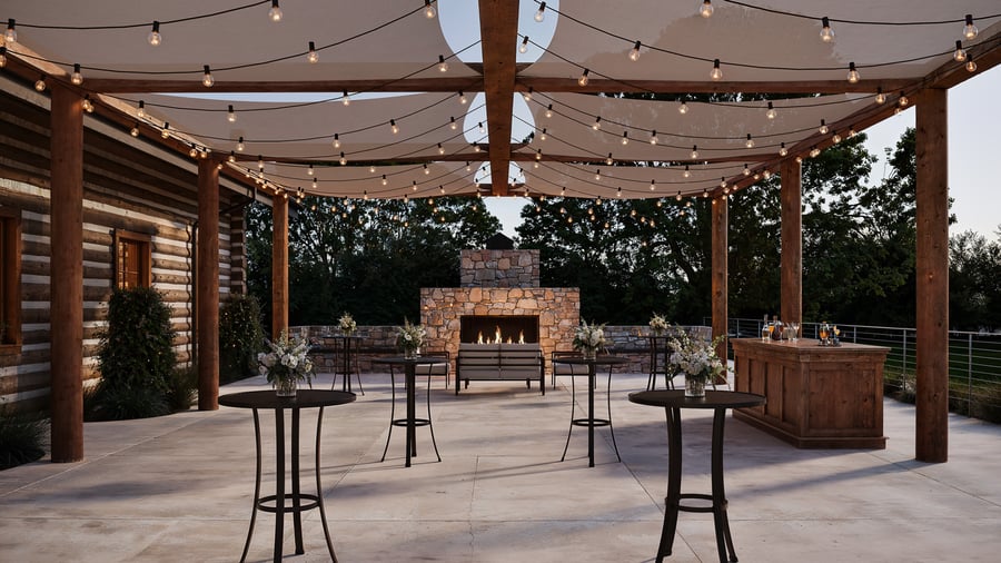 Covered outdoor patio at Casa del Rio Fresno with string lights, rustic wooden posts, stone fireplace, high-top cocktail tables with floral arrangements, wooden bar, and log cabin exterior wall creating an elegant space for outdoor gatherings.