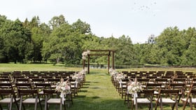 Outdoor ceremony space at Casa del Rio Fresno featuring wooden chairs arranged in rows, wooden pergola decorated with floral arrangements, lush green trees, serene pond, and natural landscape backdrop for sophisticated outdoor events.