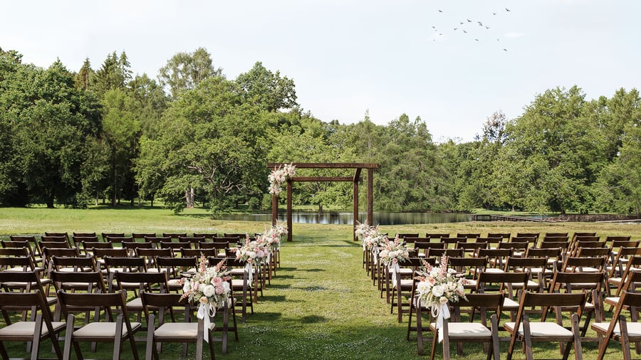 Outdoor ceremony space at Casa del Rio Fresno featuring wooden chairs arranged in rows, wooden pergola decorated with floral arrangements, lush green trees, serene pond, and natural landscape backdrop for sophisticated outdoor events.