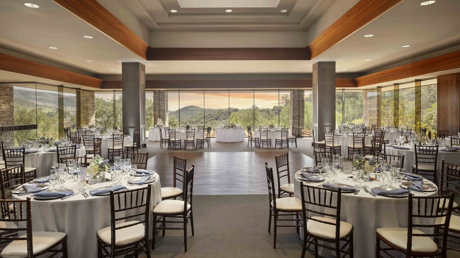 Spectacular main ballroom featuring soaring coffered ceilings with rich wood accents, floor-to-ceiling windows overlooking pristine mountain vistas, and elegant wedding reception setup with pristine white linens and mahogany chiavari chairs
