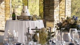 Elegant wedding cake display on silver pedestal, perfectly positioned to capture golden afternoon light streaming through floor-to-ceiling windows, with champagne flutes ready for celebratory toasts