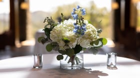 Exquisite floral centerpiece combining white hydrangeas, delicate blue delphiniums, and fresh greenery in a glass vessel, creating an elegant accent for wedding reception tables with a touch of 'something blue