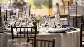 Immaculately set reception table featuring premium crystal glassware, crisp white linens, steel blue napkins, and fresh floral centerpiece, with soft natural light creating a refined dining atmosphere for wedding celebrations