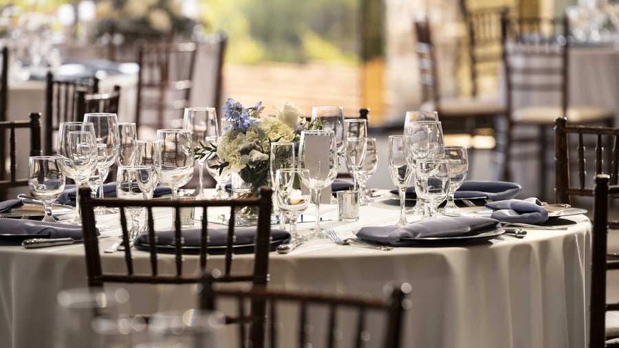 Immaculately set reception table featuring premium crystal glassware, crisp white linens, steel blue napkins, and fresh floral centerpiece, with soft natural light creating a refined dining atmosphere for wedding celebrations