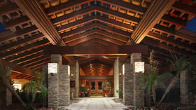 Dramatic resort entrance with soaring exposed beam cathedral ceiling, illuminated stacked stone pillars, and impressive wood double doors, creating a grand welcome for wedding guests against a backdrop of evening skies