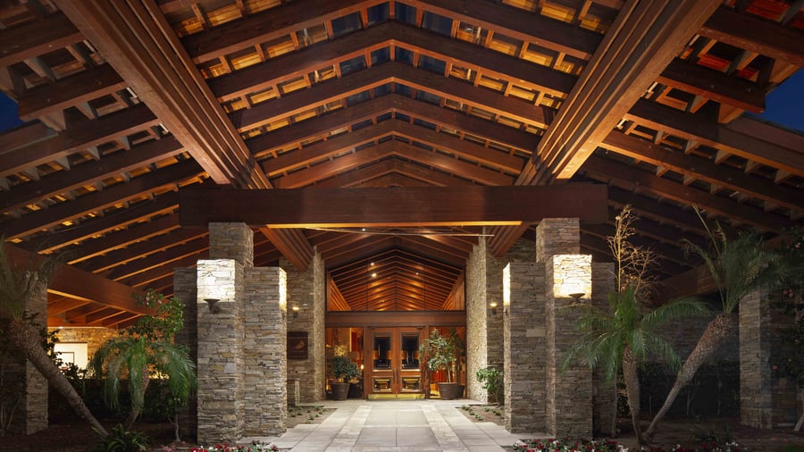 Dramatic resort entrance with soaring exposed beam cathedral ceiling, illuminated stacked stone pillars, and impressive wood double doors, creating a grand welcome for wedding guests against a backdrop of evening skies