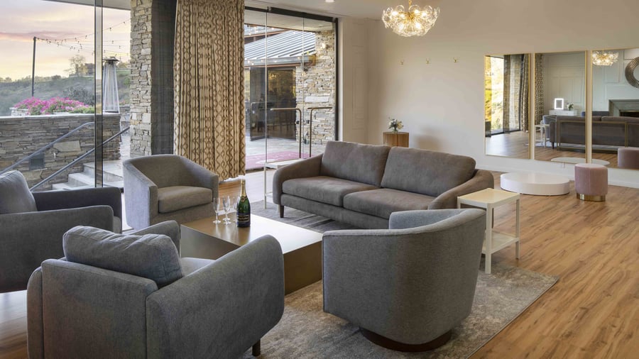 Contemporary pre-function lounge with floor-to-ceiling glass doors, designer furniture in sophisticated neutral tones, and sweeping sunset views of the surrounding hillsides
