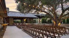 Picturesque outdoor ceremony venue with ambient string lights, Mediterranean-inspired colonnade, and rows of mahogany chairs leading to a floral-adorned focal point at sunset, creating an unforgettable ceremony backdrop