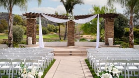 Elegant stone archway event ceremony at Fallbrook Estate by Wedgewood Events featuring white draping, white folding chairs, floral arrangements and scenic golf course backdrop for outdoor celebrations in Fallbrook California