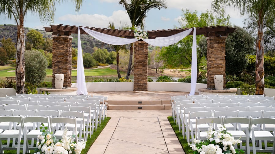 Elegant stone archway event ceremony at Fallbrook Estate by Wedgewood Events featuring white draping, white folding chairs, floral arrangements and scenic golf course backdrop for outdoor celebrations in Fallbrook California