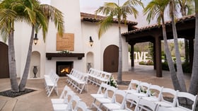 Romantic courtyard event ceremony at Fallbrook Estate featuring outdoor fireplace, palm trees, white folding chairs, signage and Spanish colonial architecture for private celebrations in Fallbrook California