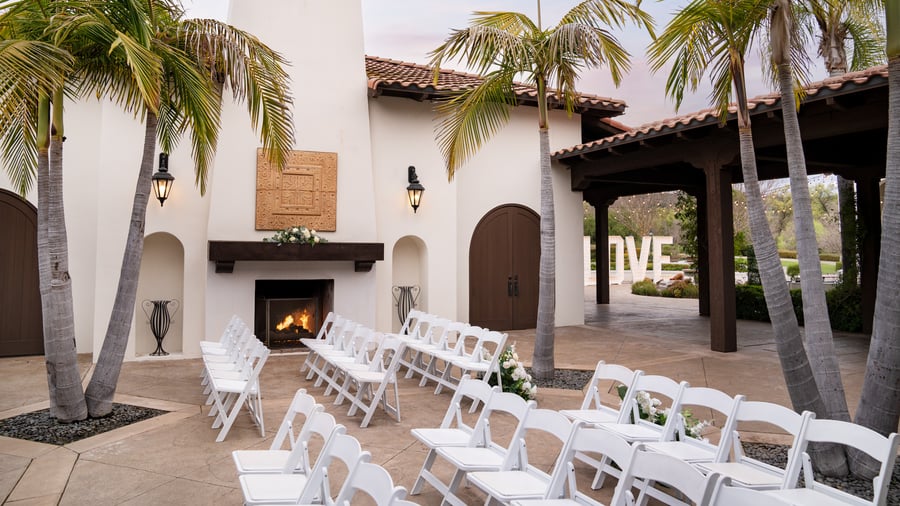 Romantic courtyard event ceremony at Fallbrook Estate featuring outdoor fireplace, palm trees, white folding chairs, signage and Spanish colonial architecture for private celebrations in Fallbrook California