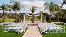 Elegant outdoor event ceremony setup at Fallbrook Estate by Wedgewood Events in Fallbrook California featuring white chairs, stone archway with flowing fabric, palm trees and golf course views for corporate events and celebrations