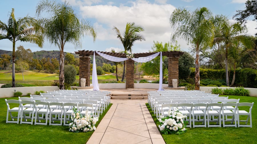 Elegant outdoor event ceremony setup at Fallbrook Estate by Wedgewood Events in Fallbrook California featuring white chairs, stone archway with flowing fabric, palm trees and golf course views for corporate events and celebrations