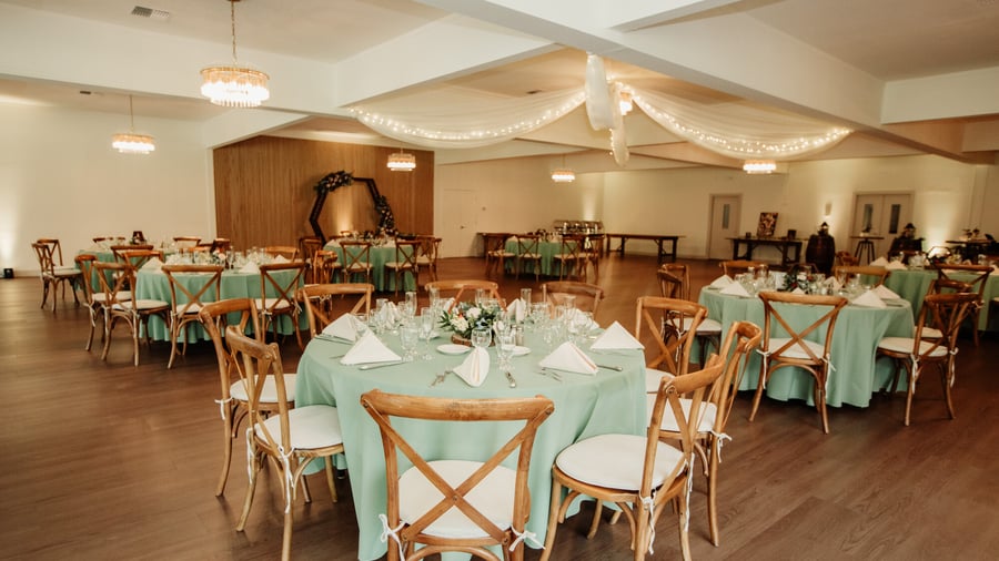 Indoor wedding reception hall at Redwood Canyon with round tables covered in mint green tablecloths, wooden cross-back chairs, white ceiling drapes, and elegant chandeliers.