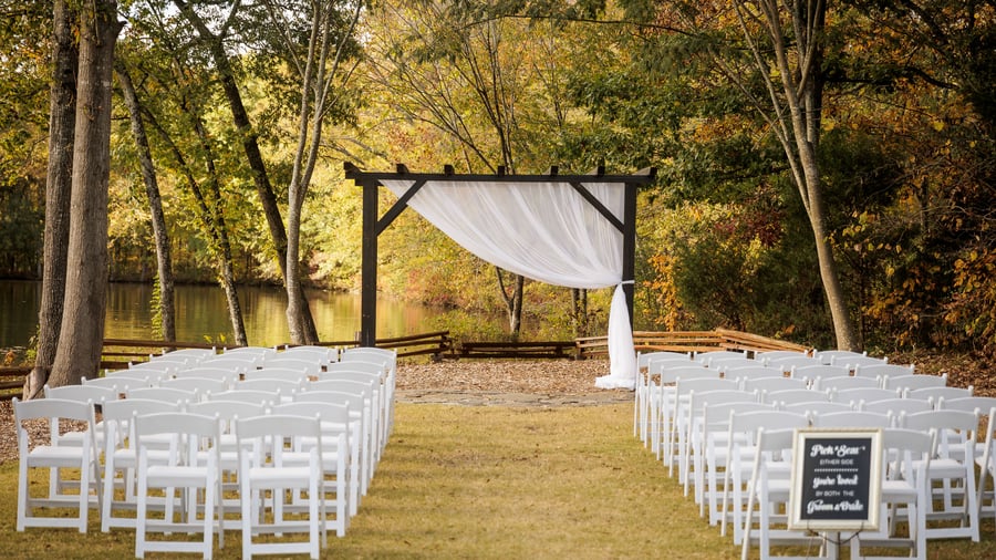Ceremony or presentation area at Riverwood Manor featuring rows of white chairs facing a wooden arbor with the tranquil lake and fall foliage as a backdrop, ideal for corporate retreats and special events - Ken Thomas Photography