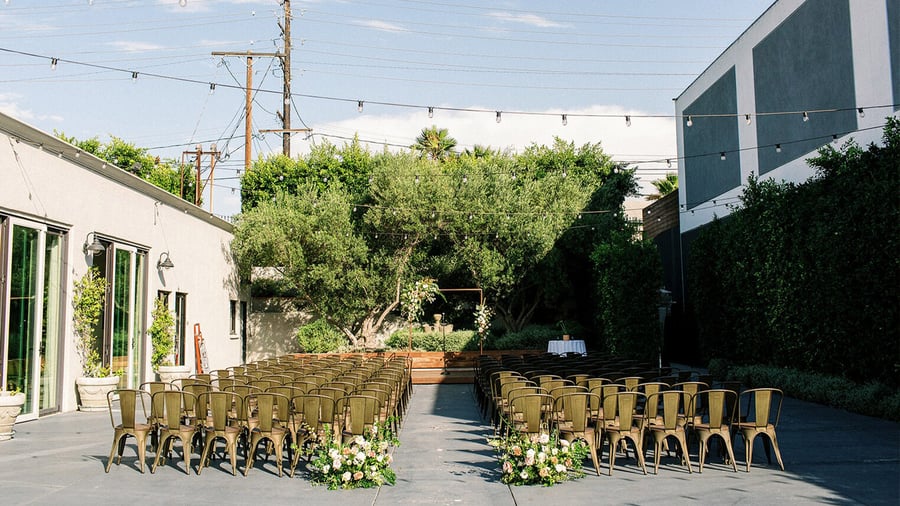 Sun-drenched courtyard ceremony space at The Harper with string lights, olive trees, and metal chairs in champagne finish